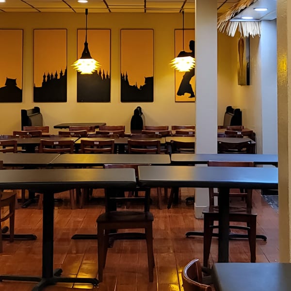 Empty dining room with dark tables and chairs, illuminated by hanging lights and wall art.