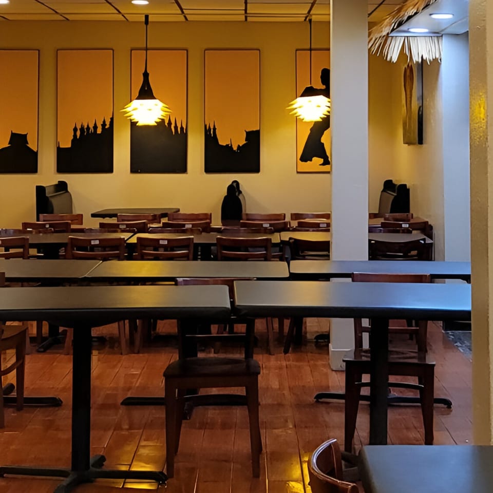 Empty dining room with dark tables and chairs, illuminated by hanging lights and wall art.