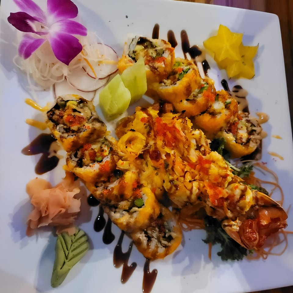 A white plate holds fried sushi rolls, a lobster tail, star fruit, and a purple orchid.