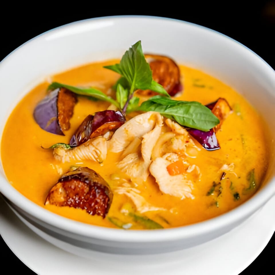 Close-up of a white bowl filled with vibrant orange Thai curry, chicken, eggplant, and basil.