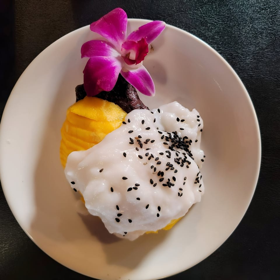 Mango sticky rice dessert with coconut cream, black sesame seeds, and an orchid garnish.