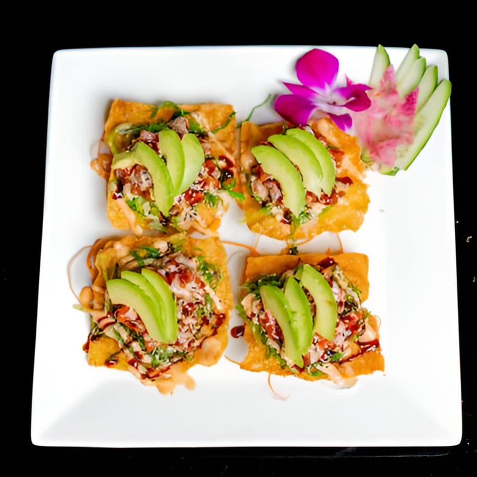 Four crispy wonton squares topped with avocado, sauce, and greens, garnished with an orchid and cucumber.